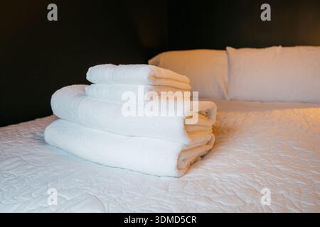 A stack of white towels on a bed. The towels are neatly folded and stacked on top of each other. The bed is made and the room is clean and tidy. Conce Stock Photo