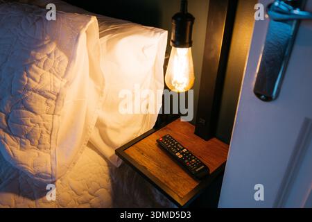 A remote control is on a wooden nightstand next to a bed. The remote control is black and silver Stock Photo