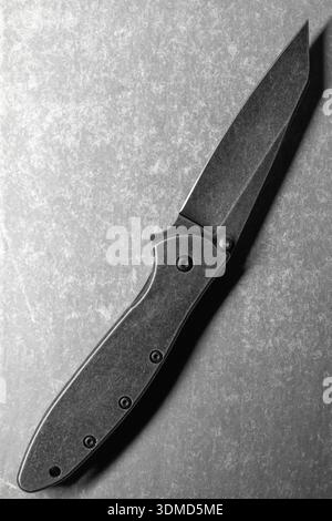 Detailed view of a modern gray stone wash steel folding knife with a sleek design lying on gray textured desk, top view Stock Photo