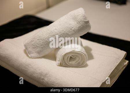 Two white towels are stacked on top of each other. The towels are folded neatly and are placed on a bed. The bed is covered with a black sheet, and th Stock Photo