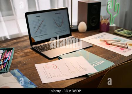 Laptop displaying math graphs on wooden desk surrounded by English workbook, graded assignment, calculator, and stationery suggesting online lesson or tutoring session setup Stock Photo