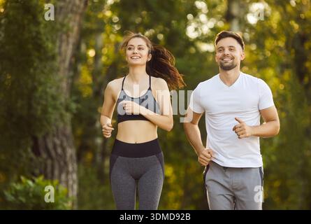 Sporty fit active man and woman running together on sunny park path smiling, green trees, happiness Stock Photo