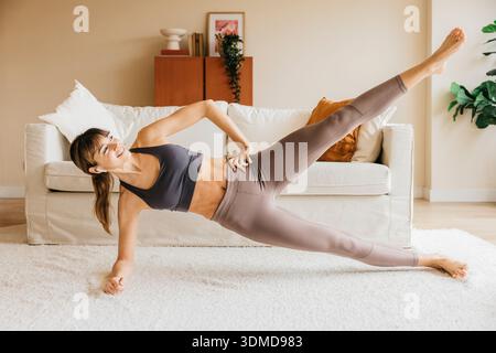 Woman performing a side plank exercise with a raised leg, smiling in her living room Stock Photo
