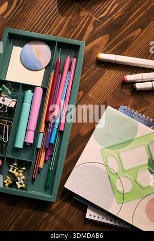 Vertical of Closeup showing organized stationery including pens, markers, eraser, paper clips, push pins, geometric stencil and spiral notebooks on wooden desk, illustrating lesson preparation Stock Photo