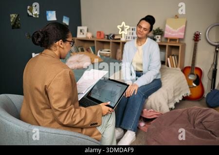 Young adult Caucasian woman sitting on bed listening attentively, while middle aged Black woman tutor explaining lesson using laptop and printed charts in cozy bedroom setting Stock Photo