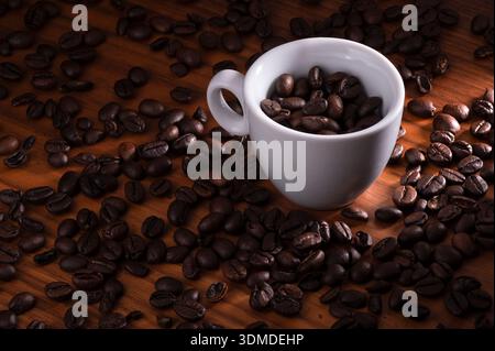 A white ceramic cup filled with dark roasted coffee beans sits on a wooden surface, surrounded by scattered beans. The warm lighting highlights the gl Stock Photo