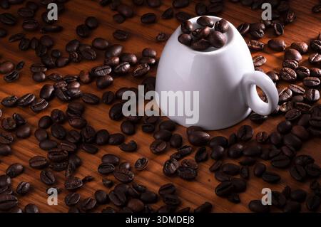 A white ceramic cup filled with dark roasted coffee beans sits on a wooden surface, surrounded by scattered beans. The warm lighting highlights the gl Stock Photo