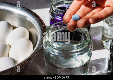 Hand holding a food dye tablet above a glass jar of water, with eggs in a bowl nearby for easter egg coloring. Stock Photo
