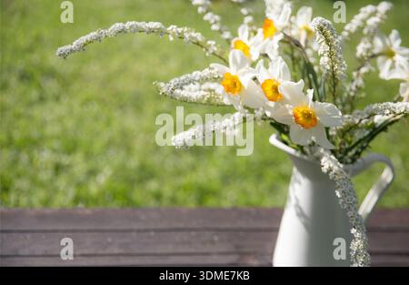 Spring still life with white daffodils and wild flowers in ceramic jug on wooden table, natural garden background, soft sunlight, copy space, seasonal Stock Photo