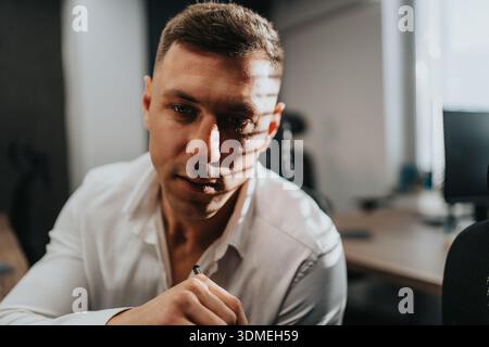 Focused man in white shirt leaning over desk with a pen in a modern office setting Stock Photo
