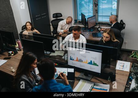 Busy office scene with professionals working at desks, monitors, and data charts in a collaborative team environment Stock Photo