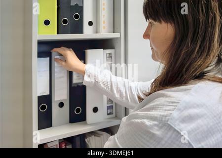 Organized continuity view of woman reaching for binders on office shelving, labeled folders and files in storage cabinet, white shirt, calm interior, Stock Photo