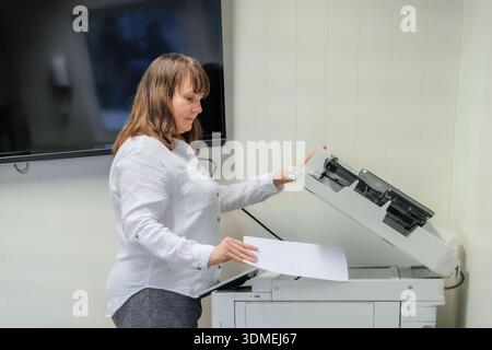 Operational flow of mature woman placing paper into large office printer or copier, side view in white shirt, neutral wall and equipment visible, rout Stock Photo
