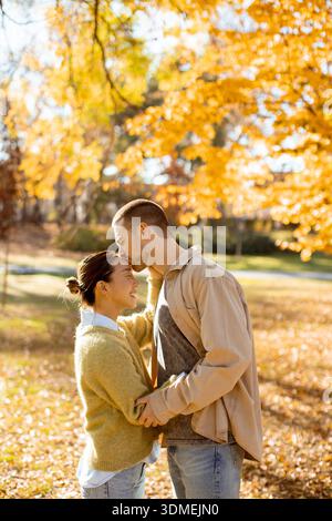 A Korean woman and a Caucasian man embrace under vibrant autumn leaves, enjoying a sunny day in the park. Their smiles radiate happiness. Stock Photo