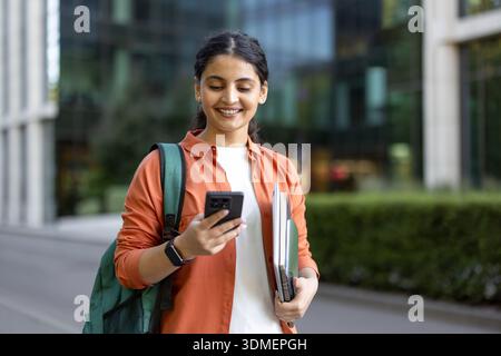 Indian woman student smiling while holding books and backpack, looking at her smartphone and walking on a modern college campus, representing education, technology, and learning Stock Photo