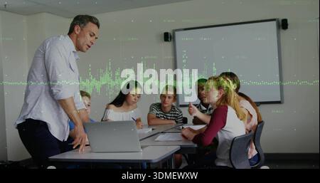 Presenting instructor in light shirt leaning over table, guiding students in classroom with laptop Stock Photo