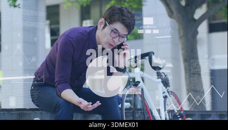 Talking mature Asian man in purple shirt holding smartphone at plaza, with bicycle and data charts Stock Photo