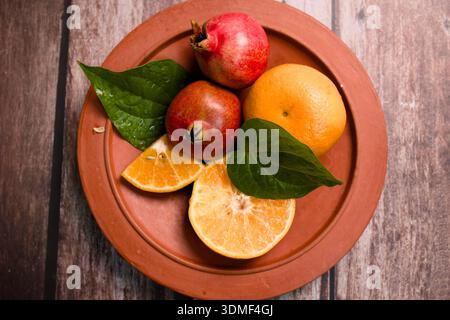 Bright fruits are arranged on a clay plate with green leaves. Two pomegranates and one orange are present. Stock Photo
