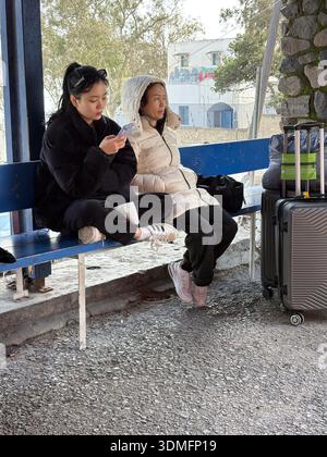 Korean mother daughter, Asian family travelers, sitting on a bench with wheeled luggage. Waiting for transportation to the airport or port. Stock Photo