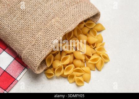 High angle shot of small Shell Pasta noodles spilling out of a burlap bag onto a light gray table. Stock Photo
