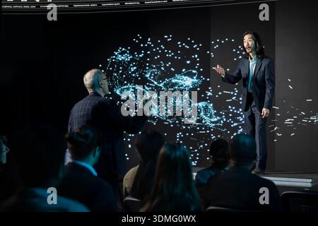 Senior businessman giving suggestions during LLM in-house debate in auditorium, analyzing AI systems as a team. Male standing up and asking questions, tech diversification. Stock Photo