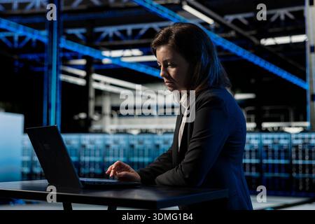Data center female programmer using laptop to inspect infrastructure, updating network systems. IT professional using notebook to ensure stable server hub operations, overseeing rigs Stock Photo