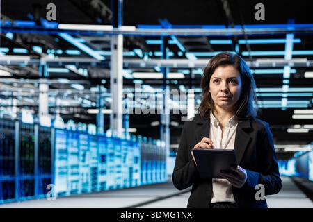 Data center technician using tablet to do checkup on cloud computing infrastructure, VFX compositing overlay. Server room employee walking between blue wireframe mainframes using device Stock Photo