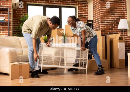 African american spouses happily carrying modern coffee table while relocating into new home. Excited married homeowners enjoying moving day, arranging furniture in fresh living room. Stock Photo