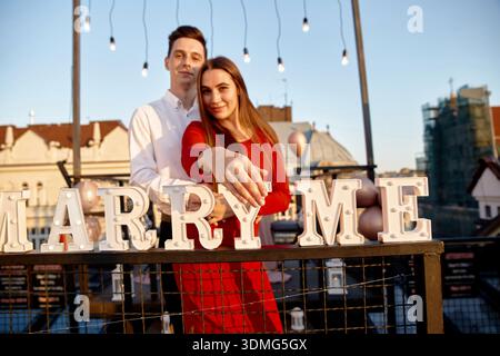 Couple stands closely on a rooftop, celebrating their engagement as they hold hands and look happy with letters displaying Marry Me. Stock Photo