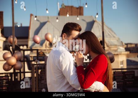 A young couple stands close together on a rooftop. They smile at each other while holding a cup. String lights hang above. Stock Photo