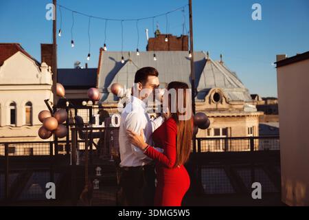 A couple stands close on a rooftop, sharing a moment together with city buildings and decorations around them during early evening. Stock Photo