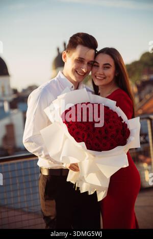 A couple stands together on a rooftop, holding a big bouquet of red roses. They smile and enjoy the evening atmosphere in the city. Stock Photo