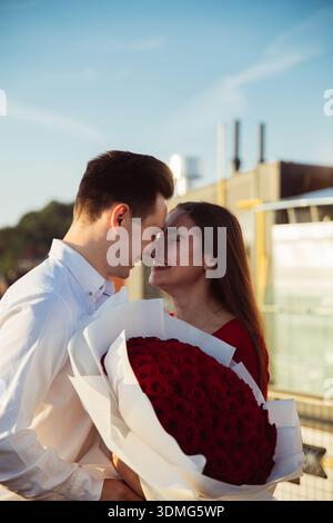 Two people hold a large bouquet of roses while smiling and leaning in for a close moment near the water during sunset. Stock Photo