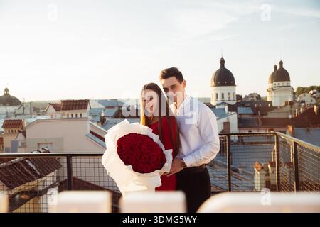 A couple stands on a rooftop holding a big bouquet of red roses while enjoying the view of the city at sunset. Stock Photo