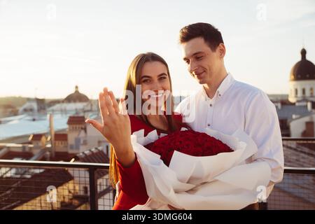 A couple stands on a rooftop with a bouquet of roses. The woman shows her ring while smiling. The sun sets over the city skyline. Stock Photo