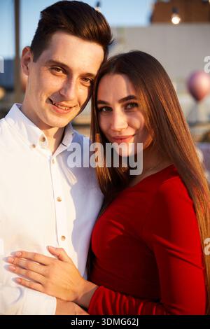 A young couple stands closely together on a rooftop during sunset with a city skyline in the background and warm light shining on them. Stock Photo