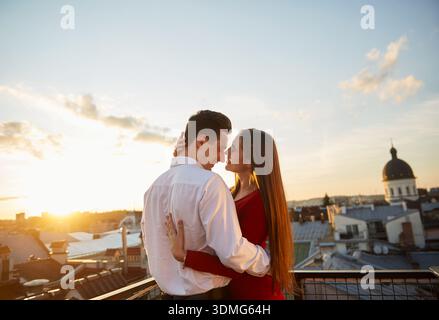 A couple stands closely on a rooftop as the sun sets. They share a moment while looking at each other against the city skyline. Stock Photo