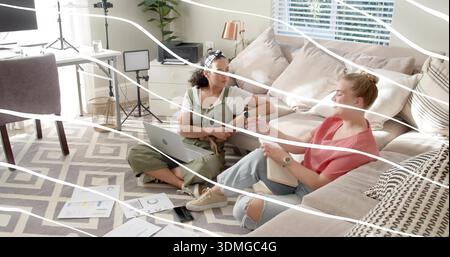 Collaborating pair of women in olive overalls, reviewing laptop, ring light, charts in living room Stock Photo