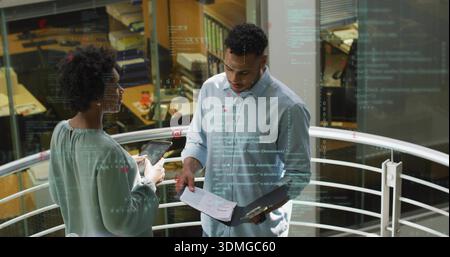 Discussing colleagues wearing blue shirt and green blouse, reviewing binder and tablet on mezzanine Stock Photo