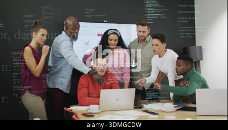 Reviewing team of seven wearing businesswear leaning over laptops at meeting table, chart display Stock Photo