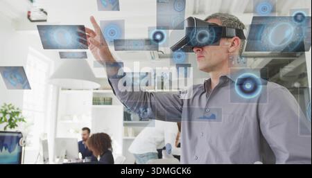 Reaching man wearing grey shirt and VR headset using holographic panels in office with monitors Stock Photo