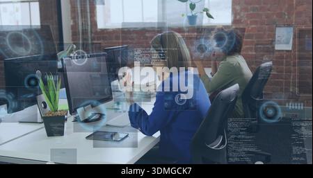 Gesturing woman in blue blazer typing and pointing at screen in loft office, with dual monitors Stock Photo