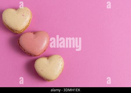 Three heart-shaped macarons are placed on a bright pink background. The macarons have different colors, with two being light beige and pink. Stock Photo