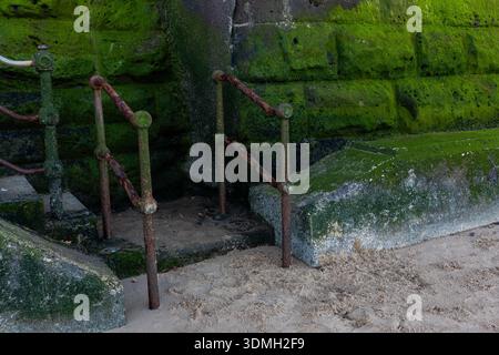 Weathered metal handrails leading to a moss covered coastal wall on a sandy beach Stock Photo
