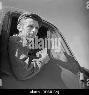 Donetsk region, Ukrainian SSR, Soviet Union, 1960s. An atmospheric black and white archival portrait of a Soviet truck driver sitting in the cabin. The young man in a flat cap leans out of the open truck window, looking into the distance with a thoughtful expression. The close-up shot captures the texture of his work clothes and the metal door frame. This vintage image documents the daily life, style, and romanticized image of the driver profession in the peaceful Donbas region during the Soviet era. Stock Photo