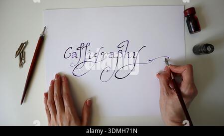A person is focusing on creating calligraphy on a blank sheet of paper. The hands hold a pen while drawing flowing letters in an elegant style. Nearby Stock Photo