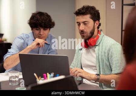 Two young male colleagues work together at a desk in a bright, modern office space, discussing a project on a laptop screen as part of a mentorship an Stock Photo