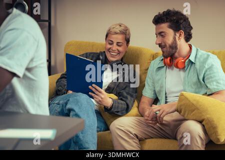 A diverse pair of young creative colleagues sit on a yellow couch in a modern startup office, laughing and discussing a project on a clipboard during Stock Photo