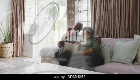 Waving man wearing grey shirt, arm around woman wearing green top, holding tablet in living room Stock Photo