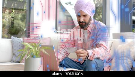 Holding card and using laptop, man in turban and plaid shirt sitting on sofa by windows Stock Photo